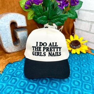May include: A baseball cap with a cream-colored crown and a black brim. The text "I DO ALL THE PRETTY GIRLS NAILS" is embroidered in black on the front. The cap is displayed with a sunflower and purple flowers.