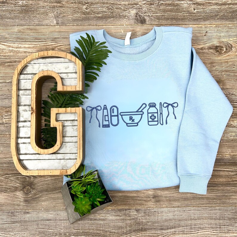a light blue sweatshirt with a graphic design on it, along with a wooden letter 'G' and a potted plant.