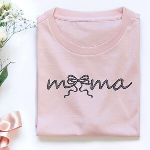 May include: A light pink t-shirt with the word "mama" in a script font, featuring a bow design in place of the letter "o". The shirt is folded neatly, with a simple crew neck design. The shirt is made of a soft material.