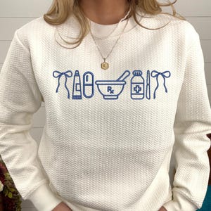 A woman wearing a white sweater with blue icons of various medical items, such as a pill bottle, a bowl, and a stethoscope, printed on it.