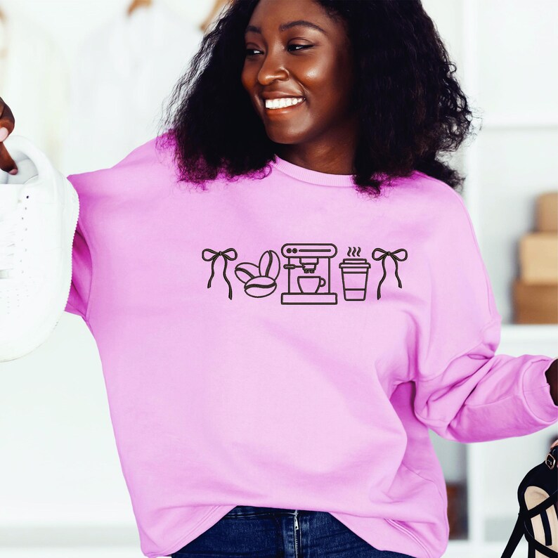 May include: A light pink sweatshirt with a black coffee-themed graphic. The graphic includes coffee beans, a coffee maker, and a to-go cup. The sweatshirt has long sleeves and a crew neck.
