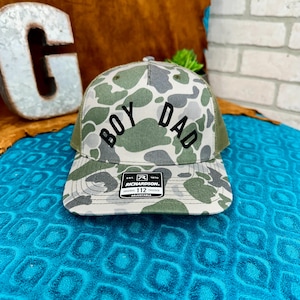 May include: A camouflage print trucker hat featuring the text BOY DAD in black lettering across the front panel. The cap includes a structured crown, a curved brim, and a black rectangular sticker on the bill with the Richardson 112 logo.