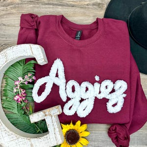 May include: A maroon crewneck sweatshirt with the word "Aggies" in white, fluffy lettering. The sweatshirt is displayed with decorative wooden letters, greenery, and a sunflower. A black hat is in the background.