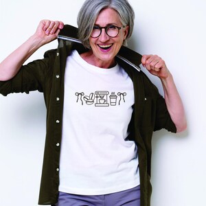 May include: A white t-shirt with a black coffee-themed graphic, including coffee beans, a coffee maker, and a to-go cup. The shirt is worn with a dark green button-up shirt and gray pants. The person is wearing glasses and smiling.