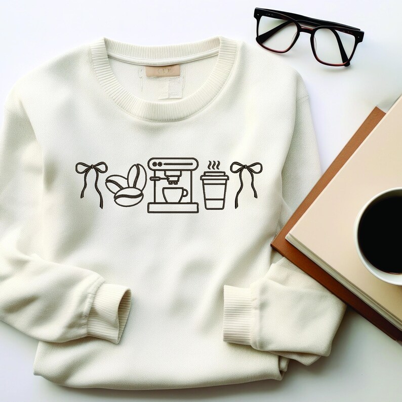 May include: Cream-colored sweatshirt with a black coffee-themed graphic. The graphic includes coffee beans, a coffee maker, a to-go cup, and bows. Black eyeglasses and a book with a coffee cup are in the background.