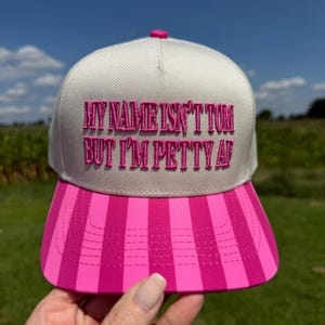 May include: A beige baseball cap with a pink striped brim. The front of the cap features embroidered hot pink text that reads, "MY NAME ISN'T TOM BUT I'M PETTY AF." The cap has a structured crown and a button on the top center of the fabric paneling.