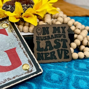 May include: A brown and black sign with the text "ON MY HUSBAND'S LAST NERVE" and a melting face graphic. The sign is surrounded by a sunflower and wooden beads. A second sign with a red letter "G" is also visible.
