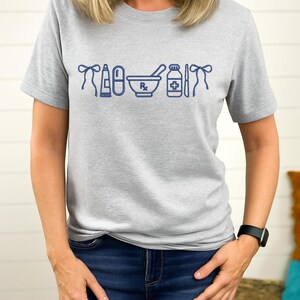 A woman wearing a gray t-shirt with a graphic design of various medical and pharmacy-related icons printed on it.
