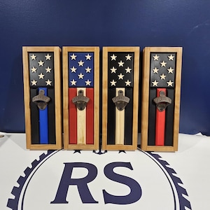 May include: Four wooden bottle openers with American flag designs. Each opener has a different color stripe: blue, red, black, and red. The top section features white stars on a black background. The openers are framed in natural wood.