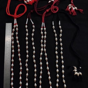 May include: Four silver bead necklaces with red, burgundy, and black cords. Each necklace has a tassel at the end of the cord. The necklaces are laid out on a black surface with a ruler for scale.