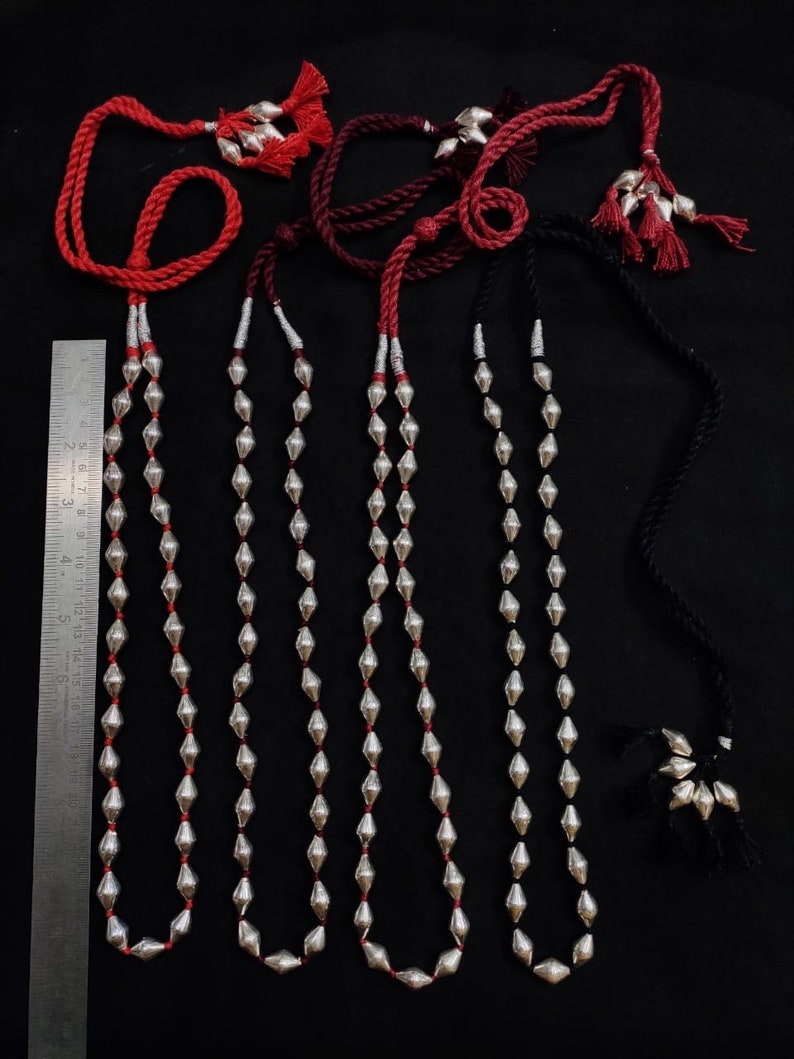May include: Four silver bead necklaces with red, burgundy, and black cords. Each necklace has a tassel at the end of the cord. The necklaces are laid out on a black surface with a ruler for scale.