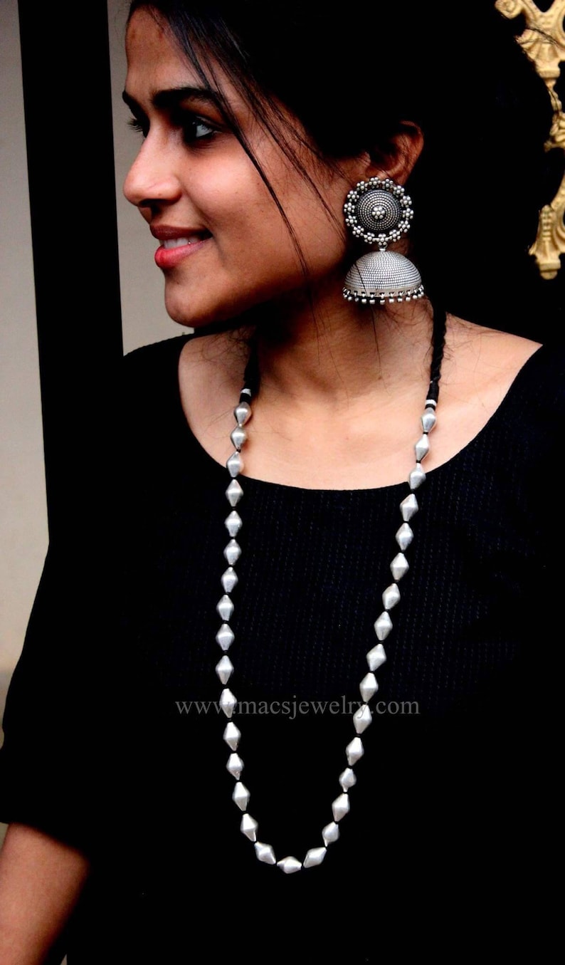 May include: A silver necklace with a long chain of diamond-shaped beads. The necklace is worn by a person with dark hair and a black shirt. The text "www.macsjewelry.com" is visible on the shirt.