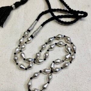 May include: A silver beaded necklace with a black cord and tassel. The beads are shaped like elongated diamonds and are strung on a black cord. The necklace has a tassel at the end of the cord.