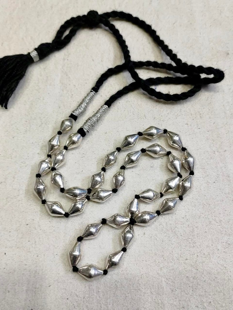May include: A silver beaded necklace with a black cord and tassel. The beads are shaped like elongated diamonds and are strung on a black cord. The necklace has a tassel at the end of the cord.