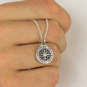Op de afbeelding: Zilveren kompas hanger ketting met de woorden "Adventure Awaits, Travel Safe" gegraveerd rond de rand. De hanger heeft een sterontwerp in het midden. De ketting wordt in een hand gehouden tegen een witte achtergrond.