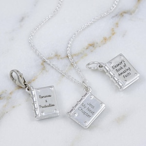 Peut inclure: Trois breloques en forme de livre argenté, dont une attachée à une chaîne. Chaque breloque présente une couverture rectangulaire avec un dos et un texte gravé : Romances & Practicalities, Jess My Next Chapter et Eleanor's Book of Amazing Facts.