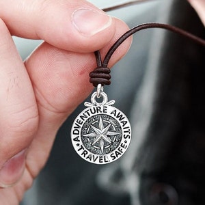 Op de afbeelding: Een zilveren kompas hanger ketting met een bruin leren koord. Het kompas heeft de tekst "Adventure Awaits Travel Safe".