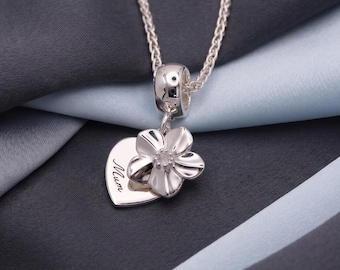 May include: A silver-toned necklace featuring a heart-shaped pendant with the word Mum engraved on it. Attached to the heart is a five-petaled flower charm with a small clear stone at its center. The pendant hangs from a delicate silver chain.