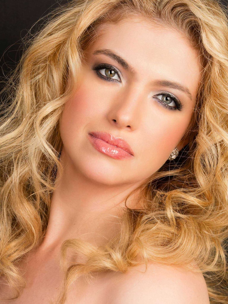 May include: A portrait of a person with long, wavy blonde hair. The person has fair skin, with makeup including eyeshadow, blush, and glossy lipstick. A small, sparkly earring is visible. The background is dark.
