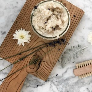 May include: A glass jar filled with a white and lavender-colored sugar scrub, sitting on a wooden cutting board with dried lavender sprigs and white flowers.