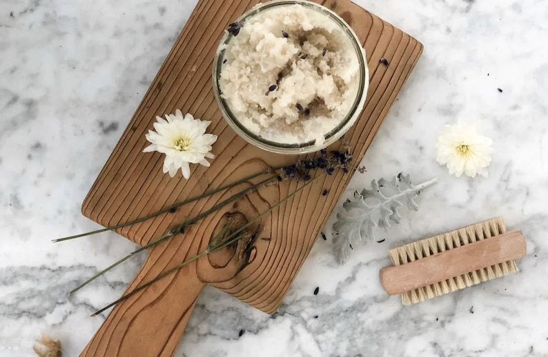 May include: A glass jar filled with a white and lavender-colored sugar scrub, sitting on a wooden cutting board with dried lavender sprigs and white flowers.