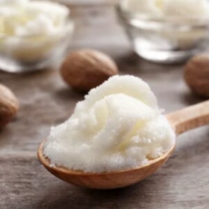 May include: A wooden spoon filled with white shea butter, surrounded by brown nutmegs on a wooden surface.