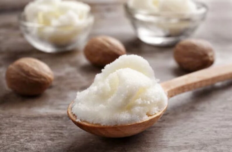 May include: A wooden spoon filled with white shea butter, surrounded by brown nutmegs on a wooden surface.