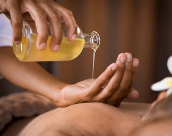 May include: A person's hand pouring clear oil from a glass bottle into another person's hand. The person receiving the oil is lying on their stomach with a white flower on their head.
