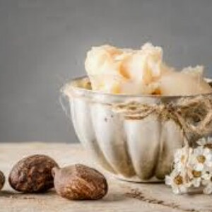 May include: A silver bowl filled with shea butter, with three shea nuts in the foreground. The bowl is tied with twine and has a decorative, fluted design.