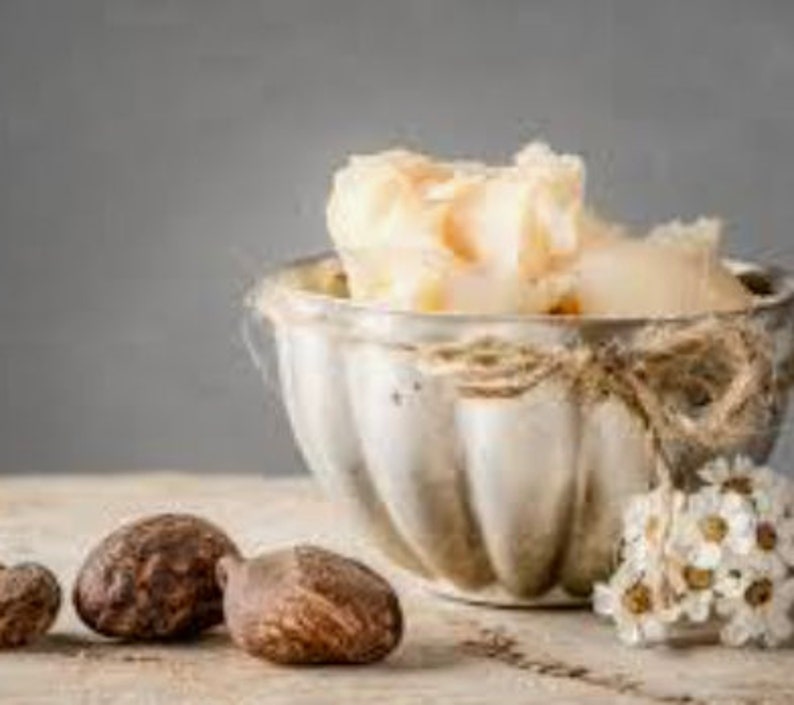 May include: A silver bowl filled with shea butter, with three shea nuts in the foreground. The bowl is tied with twine and has a decorative, fluted design.