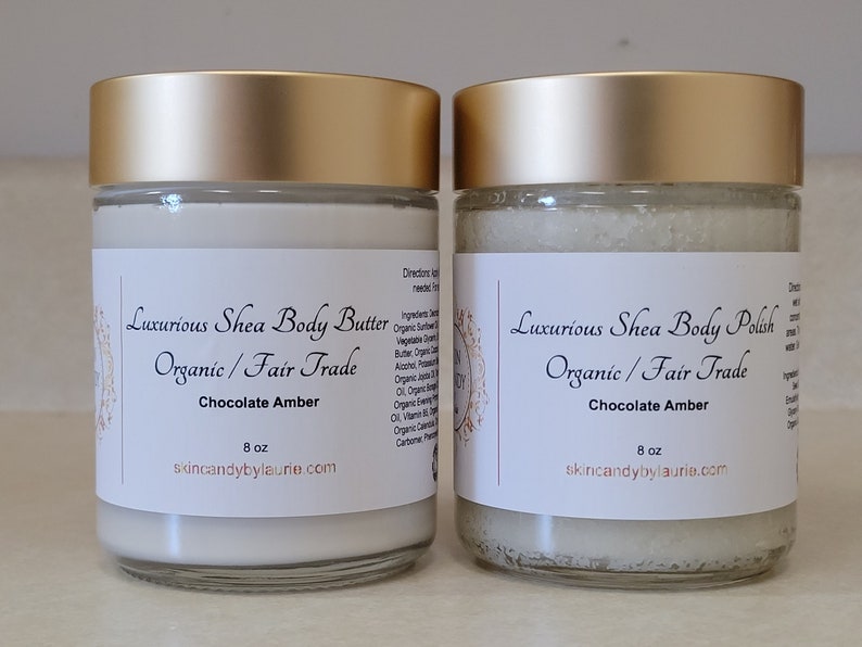 May include: Two glass jars with gold lids containing body care products. The jar on the left contains shea butter and the jar on the right contains shea body polish. Both jars are labeled "Luxurious Shea Body Butter/Polish Organic/Fair Trade Chocolate Amber 8 oz skincandybylaurie.com"