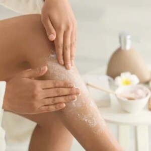 May include: A person is applying a white body scrub to their leg. The scrub is visible on the skin. The person is wearing a white robe. There is a white table in the background with a bowl of pink scrub and a bottle of lotion.