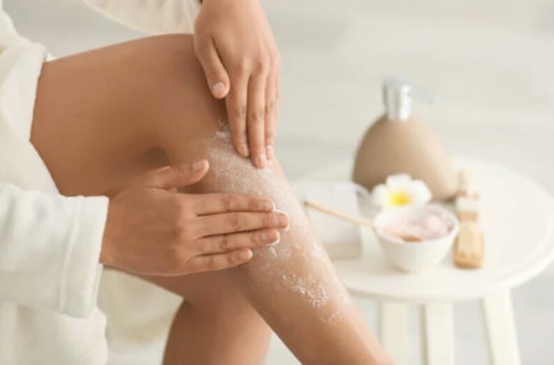 May include: A person is applying a white body scrub to their leg. The scrub is visible on the skin. The person is wearing a white robe. There is a white table in the background with a bowl of pink scrub and a bottle of lotion.