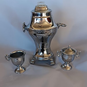 May include: A shiny, silver tea or coffee service set. The set includes a large urn with a glass top, a creamer, and a lidded sugar bowl. The pieces have ornate handles and are displayed on a light gray surface.
