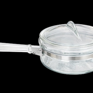 Puede incluir: Cacerola de vidrio transparente con tapa y asa. La cacerola tiene una banda de metal plateado y un asa transparente. La tapa tiene un asa de bucle. Sobre un fondo negro.
