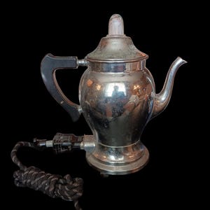 May include: Vintage chrome electric tea kettle with a black handle and a clear, ridged knob on the lid. The kettle has a spout and a black and white braided power cord with a two-prong plug.