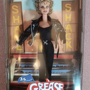 Può includere: Una bambola Barbie Collector Edition in una scatola con marchio Grease. La bambola ha capelli biondi ricci e indossa una giacca di pelle nera sopra una tuta nera con fibbia argentata. La confezione riporta le scritte 25 years e Grease.