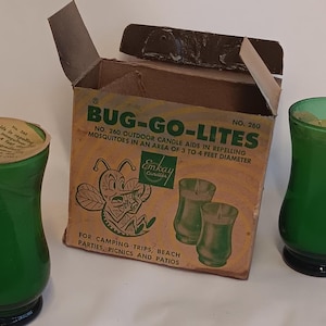 Op de afbeelding: Een doos Bug-Go-Lites buitenkaarsen, met het label "No. 260 Outdoor Candle Aids in Repelling Mosquitoes in an area of 3 to 4 feet diameter." De doos heeft een cartoon-bij en het Emkay Candles-logo. Twee groene glazen kaarsenhouders zijn te zien.