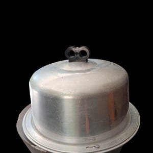 May include: A vintage, silver-colored cake carrier with a domed lid and a decorative handle. The carrier has a circular base and a matching lid, perfect for transporting or displaying baked goods. The handle is a dark color and has a unique design.