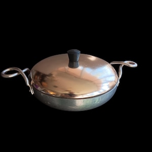 May include: A round, silver-toned metal cooking pot with a matching lid and black handle. The pot has two curved handles on either side. The lid is slightly domed and reflects the surrounding environment. The pot is set against a black background.