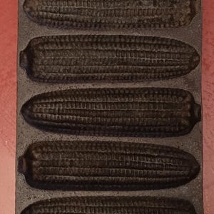 Puede incluir: Un molde para pan de maíz de hierro fundido con seis moldes individuales con forma de mazorcas de maíz. El molde es negro y tiene un asa en la parte superior.