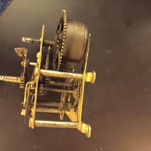 May include: A vintage clock mechanism featuring brass-coloured gears, wheels, and supporting structures. The intricate internal workings of the clock are visible, showcasing the mechanical components. The clock mechanism is set against a dark background.