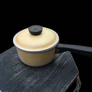 Puede incluir: Una pequeña cacerola amarilla vintage con un asa y tapa negras. La tapa tiene un pomo oscuro. La cacerola se encuentra sobre una superficie oscura y texturizada sobre un fondo negro. La cacerola es probablemente de metal.