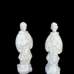 May include: Two white ceramic figurines depicting standing figures in traditional robes. The left figure wears a head covering and holds a small pouch. The right figure wears an ornate headdress and holds a small circular object with a central relief.