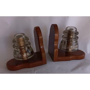 Op de afbeelding: Een paar houten boekensteunen met elk een heldere glazen isolator. De boekensteunen zijn gemaakt van donkerbruin hout en hebben een gebogen ontwerp. De glazen isolatoren zijn helder en hebben een unieke vorm, wat een vintage uitstraling geeft.