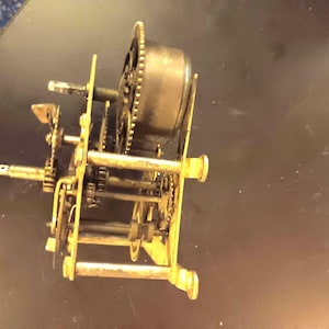 May include: A mechanical clock movement assembly featuring a series of brass plates, interconnected metal gears, and a cylindrical mainspring barrel. The internal mechanism is exposed, showing various shafts, pinions, and small metal components.