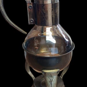 May include: A silver and glass coffee pot with a decorative floral design on the side. The pot has a glass carafe and a metal stand with three legs.