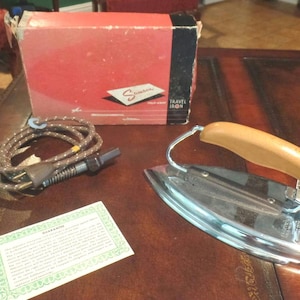 May include: A vintage Sunbeam Travel Iron in a silver and chrome finish with a brown handle. The iron is in its original box with a warranty card.