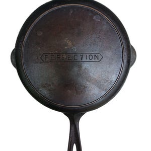 May include: A vintage cast iron skillet with a single handle and two small side handles. The word "PERFECTION" is embossed on the base of the pan. The pan is dark brown with signs of wear.
