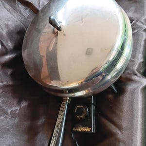 Può includere: Una padella elettrica rotonda in acciaio inossidabile Farberware con coperchio lucido dotato di manico nero. L'apparecchio include un manico nero con il marchio Farberware e un'unità di controllo del calore nera rimovibile con manopola e cavo.